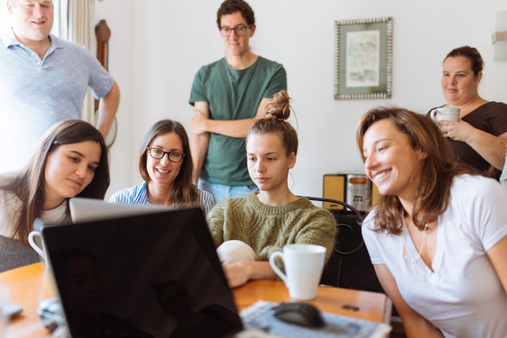 Sieben junge Menschen sitzen und stehen vor einem Laptop, auf den sie gemeinsam schauen. 